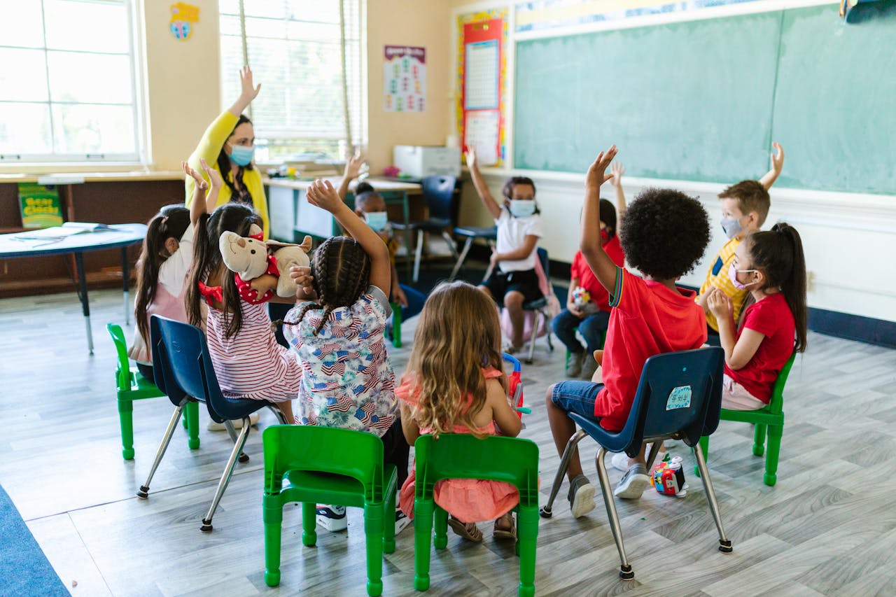 Diverse group of children in a classroom, raising hands, led by masked teacher. Bright and engaging learning environment.